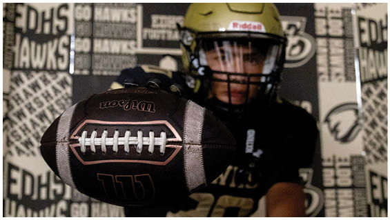 A person wearing a gold football helmet and black clothing is holding a football close to the camera with a background covered in sports-themed logos and text. Description generated by AI