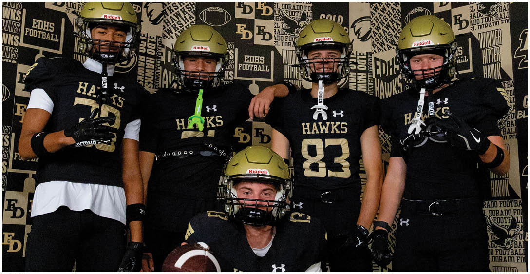 Five people wearing black football uniforms with gold helmets and the word “HAWKS" pose together in front of a backdrop with repeated text. Description generated by AI