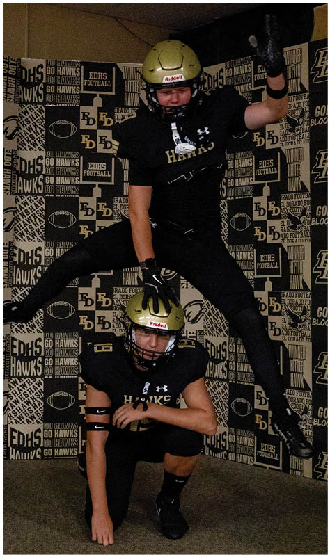 Two people in black football uniforms and gold helmets pose with one person crouching and the other jumping over them with one arm raised. Description generated by AI