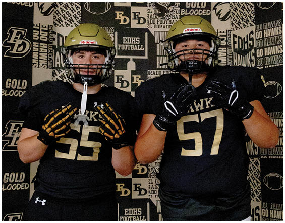 Two people wearing black football uniforms and gold helmets pose in front of a backdrop covered with football-related logos and text. Description generated by AI