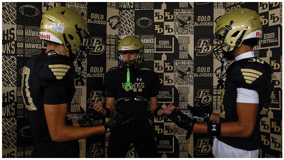 Three people wearing sports gloves and athletic clothing stand facing each other with their hands extended in front of a backdrop covered in sports-related logos and text. Description generated by AI