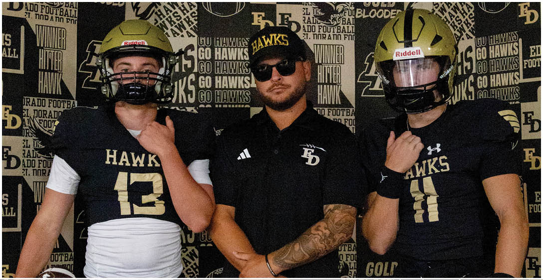 Three people wearing football helmets and black sports jerseys with the word “HAWKS" pose in front of a backdrop covered with sports-related text and logos. Description generated by AI