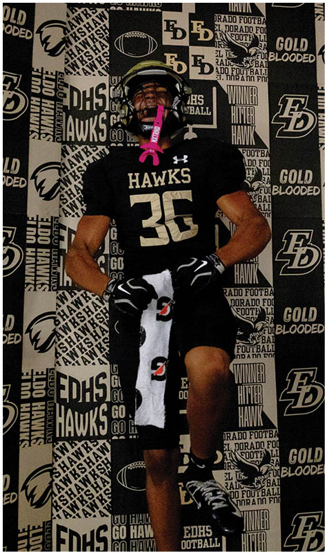 A person wearing a black football uniform with the number 36 and the word Hawks stands in front of a backdrop covered with various sports-related logos and text. Description generated by AI