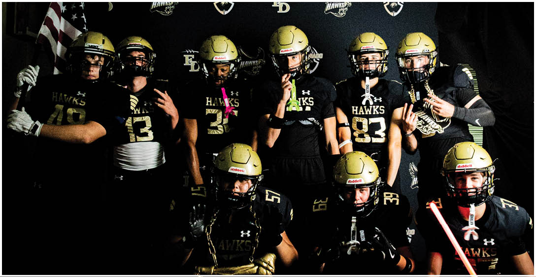 A group of people wearing black and gold football uniforms and helmets pose together in front of a backdrop with logos and an American flag. Description generated by AI