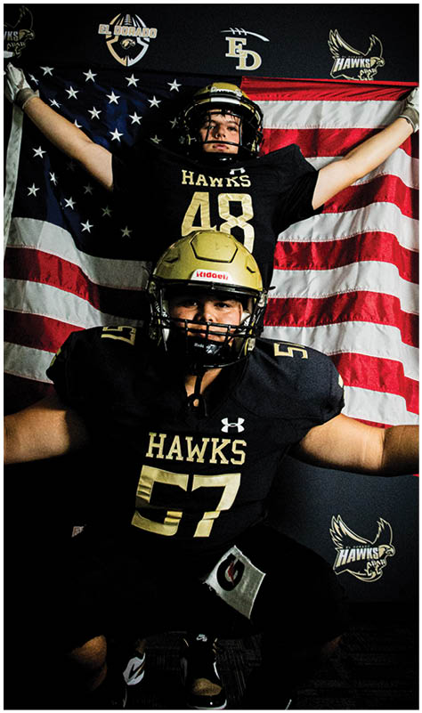 Two people wearing black football uniforms with gold helmets and the word “HAWKS" pose in front of an American flag, with one person standing behind the other and holding the flag outstretched. Description generated by AI