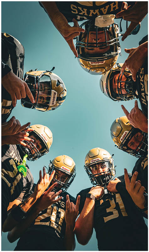A group of people wearing football helmets and jerseys are standing in a circle looking down toward the camera with their hands raised in various gestures against a clear blue sky. Description generated by AI