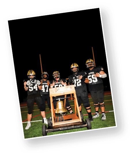 Five people in football uniforms stand on a field at night around a large bell mounted on a wooden frame with wheels. Description generated by AI