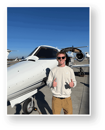 A person wearing sunglasses, a light-colored sweatshirt, and tan pants is standing on a sunny tarmac giving two thumbs up in front of a small white private jet. Description generated by AI