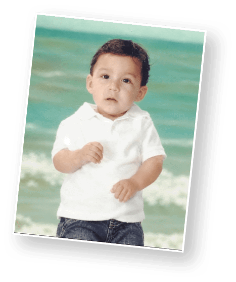 A child with short curly hair wearing a white polo shirt and blue jeans stands in front of a blurred ocean background. Description generated by AI