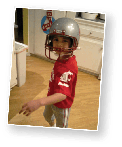 A child wearing a red sports shirt and a silver football helmet is standing in a kitchen with light wood flooring and white cabinets. Description generated by AI