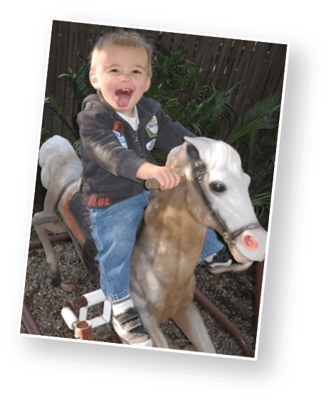A child with short curly hair wearing a white polo shirt and blue jeans stands in front of a blurred ocean background. Description generated by AI