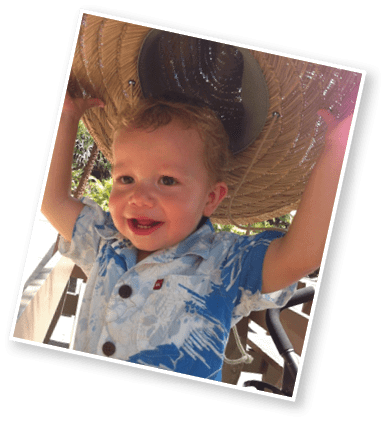 A child with short curly hair wearing a white polo shirt and blue jeans stands in front of a blurred ocean background. Description generated by AI