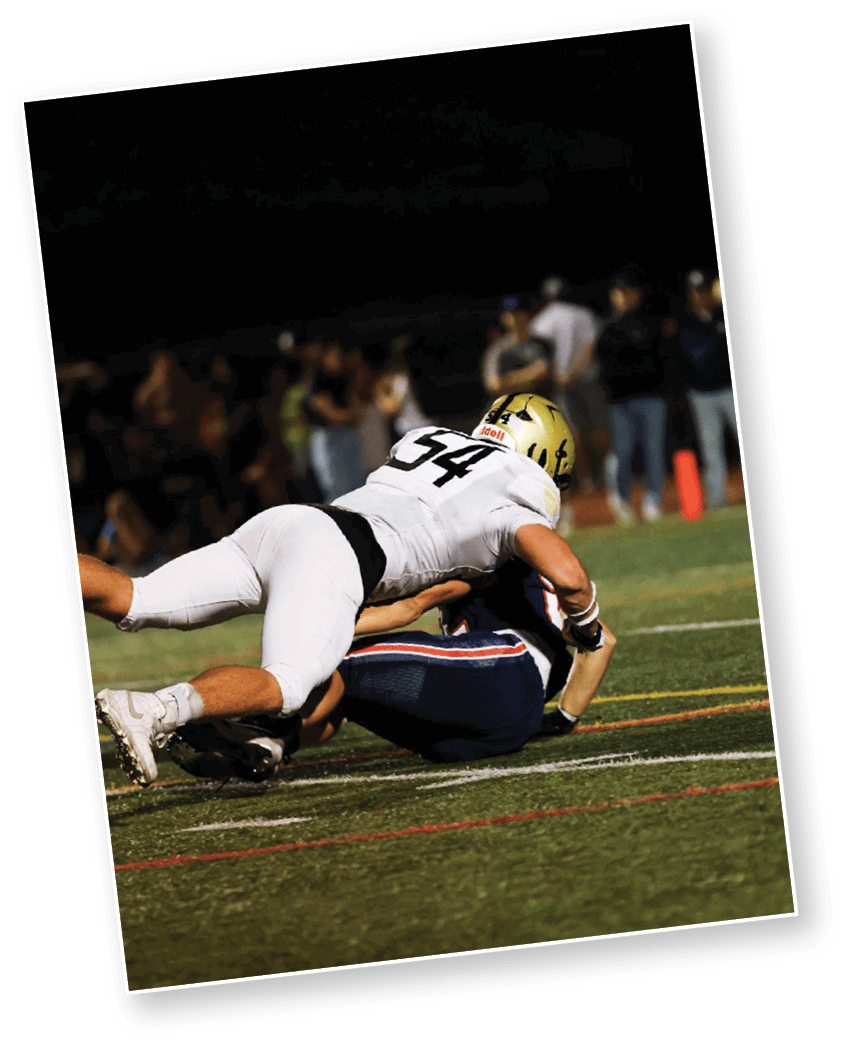 A person wearing a white football uniform with the number 54 is tackling another person in a dark blue uniform on a football field at night. Description generated by AI