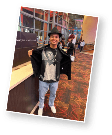 A person wearing a hat and a jacket open to reveal a t-shirt with a graphic of a smiling person in a tuxedo stands inside a brightly lit building with large windows and a carpeted floor. Description generated by AI