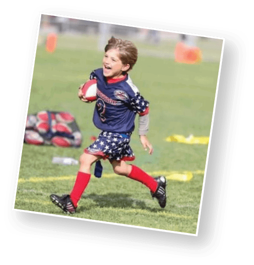 A child wearing a star-spangled sports uniform and red socks is running joyfully on a grassy field while holding a football. Description generated by AI