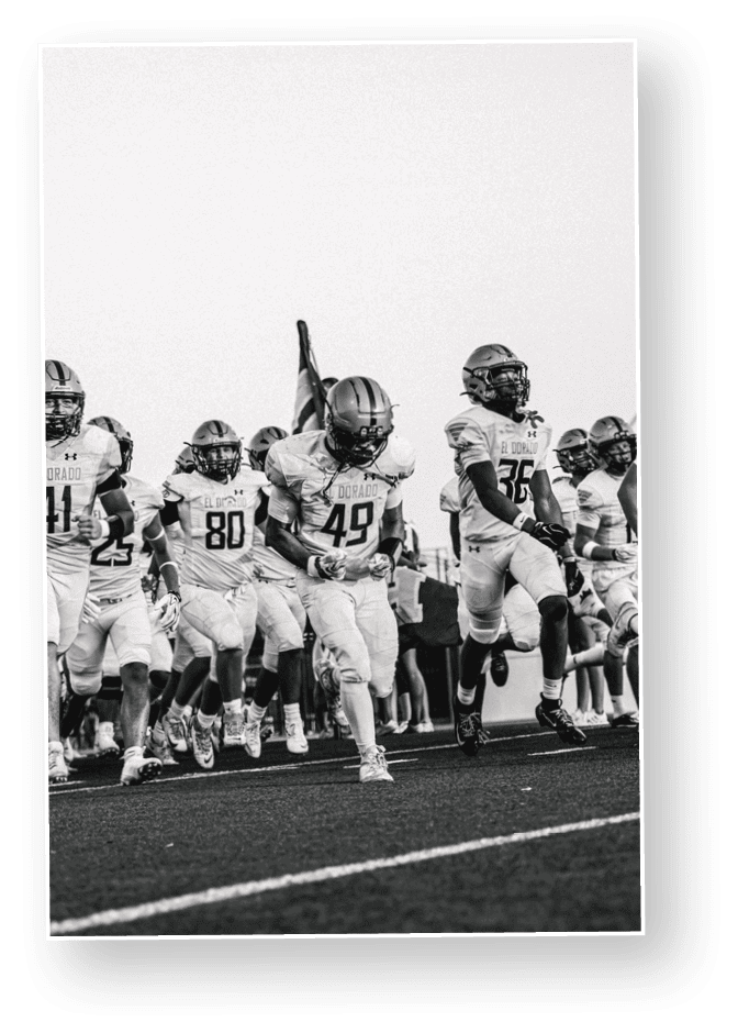 A group of people wearing football uniforms and helmets are running forward on a field with one person holding a flag. Description generated by AI