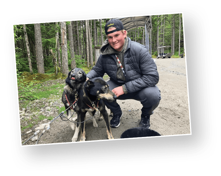 A person wearing a black cap and jacket is crouching on a dirt path in a forest while holding two black and tan dogs on leashes. Description generated by AI