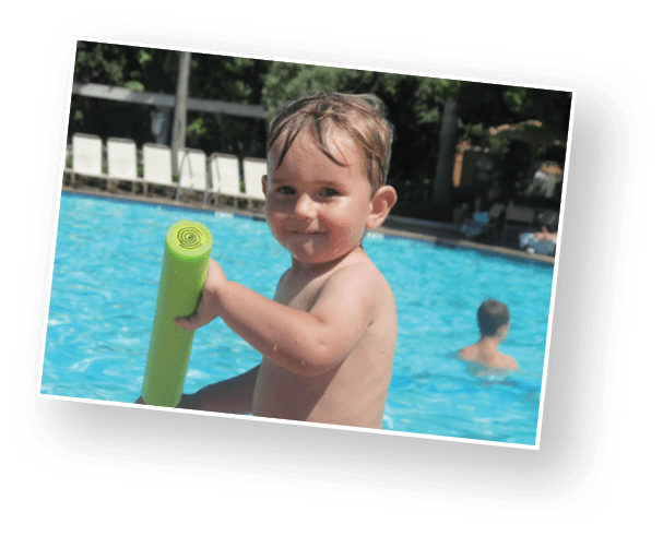 A child with wet hair is holding a green pool noodle while sitting at the edge of a swimming pool with another person in the water in the background. Description generated by AI