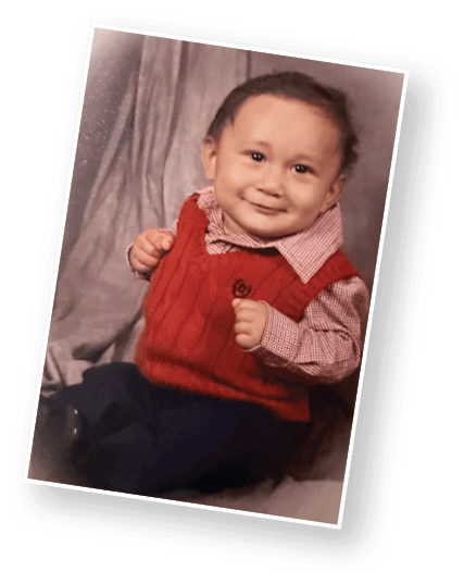A child wearing a red knitted vest over a checkered shirt is sitting with clenched fists and a slight smile. Description generated by AI