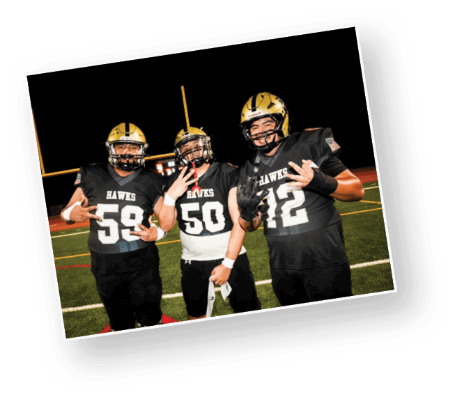 Three people wearing black football uniforms with gold helmets stand on a field at night, posing with hand gestures and smiling. Description generated by AI