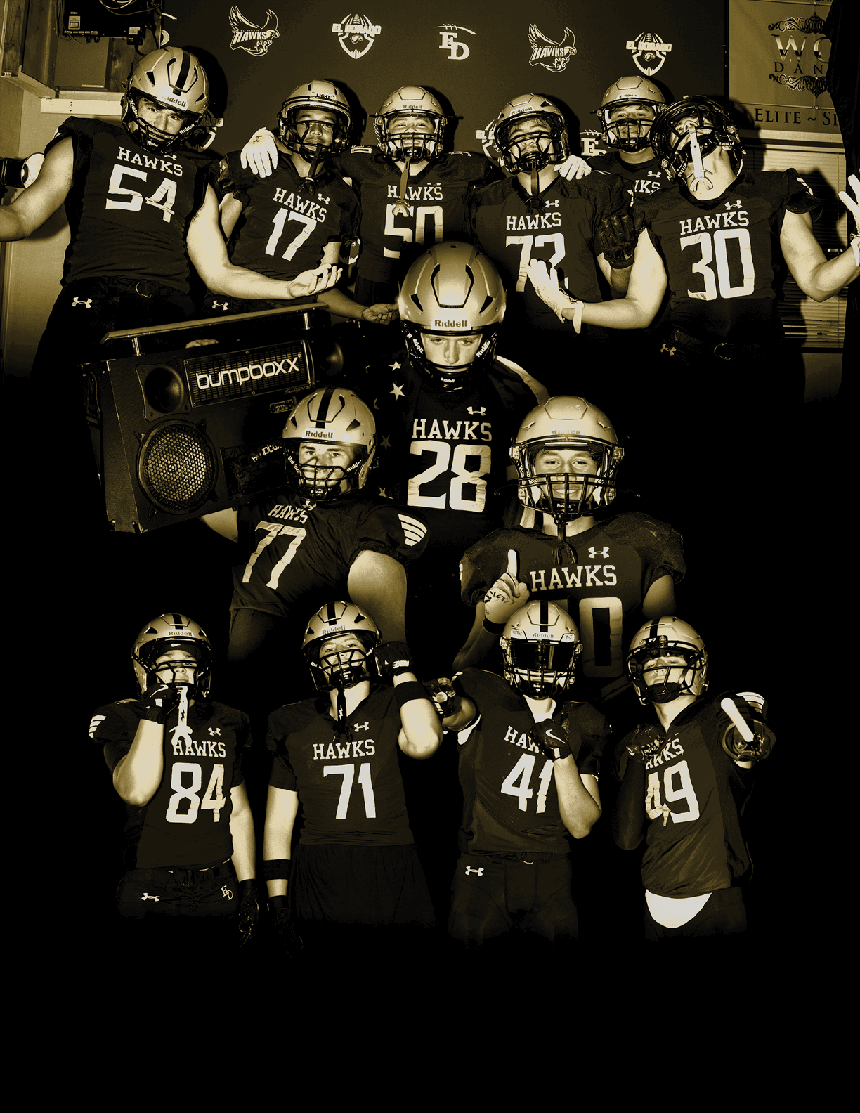 A group of people wearing football helmets and jerseys with the word “HAWKS" pose together in a dimly lit room with a speaker and a backdrop featuring logos. Description generated by AI