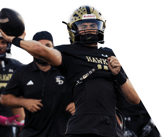 A person in a black and gold football uniform is throwing a football while several teammates in similar uniforms stand in the background on a field. Description generated by AI