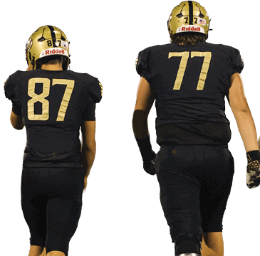 Four people wearing black football uniforms and gold helmets walk side by side on a football field at dusk. Description generated by AI
