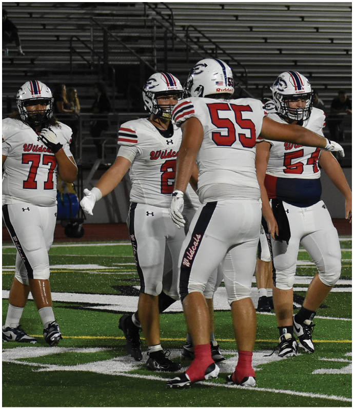 A group of people wearing white football uniforms with helmets are standing and interacting on a football field with empty bleachers in the background. Description generated by AI