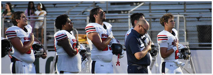 Several people in football uniforms and one person in a polo shirt stand side by side on a field with their hands over their hearts, facing the same direction. Description generated by AI