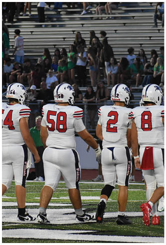 Two people wearing white football uniforms with red numbers 58 and 2 stand on a field with spectators in the background. Description generated by AI