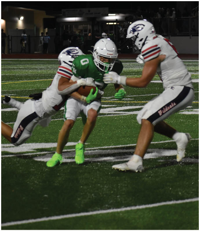 Three people in football uniforms are actively engaged in a play on a well-lit field at night with a referee observing in the background. Description generated by AI