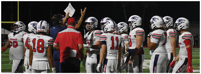 A group of football players in white uniforms with helmets are gathered around a coach wearing a red shirt on a field at night. Description generated by AI