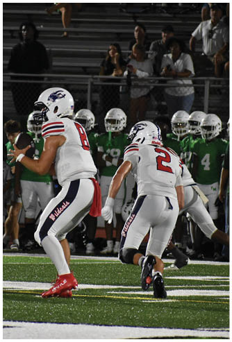 A person in a white football uniform with “Wildcats" on the pants is holding a football and preparing to throw while another person in a similar uniform is nearby on a football field with people in green uniforms in the background. Description generated by AI