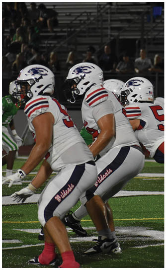 Three people in white football uniforms and helmets are positioned on a field, appearing ready to start a play during a nighttime game. Description generated by AI