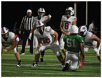 Several football players in white and green uniforms are positioned on a field during a nighttime game with a referee and other people visible in the background. Description generated by AI