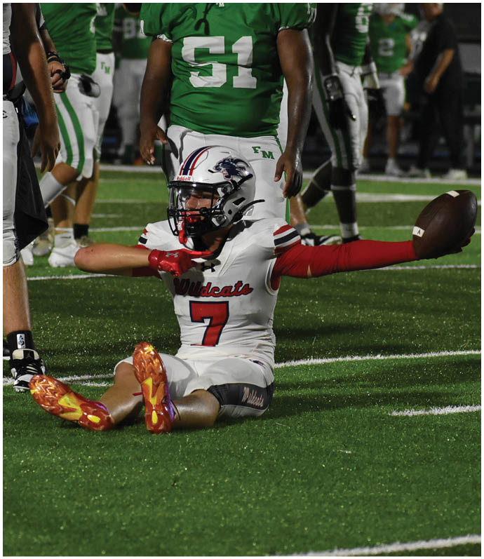 A person wearing a white football uniform with the number seven is sitting on the field holding a football while other players in green and white uniforms stand nearby. Description generated by AI