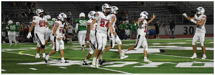 People in white football uniforms are walking on a field with green and yellow markings while people in green uniforms are in the background near bleachers. Description generated by AI