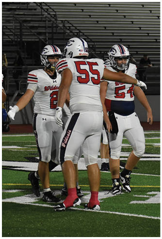 Three people in white football uniforms with helmets stand on a field, with one person wearing a jersey numbered 55 and pointing. Description generated by AI