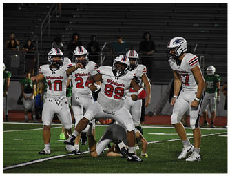 Five people in white football uniforms with helmets are on a field, with one person in the center striking a celebratory pose while others stand nearby. Description generated by AI