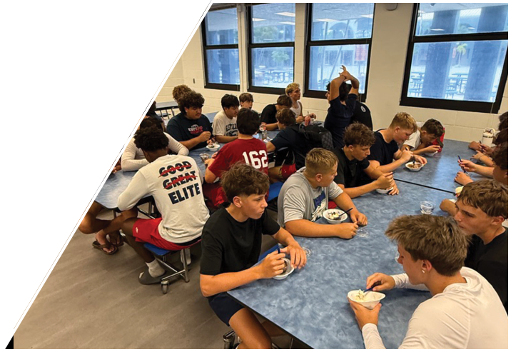 A group of people sitting closely together in a room with large windows, some engaged in conversation and one person raising their hands. Description generated by AI