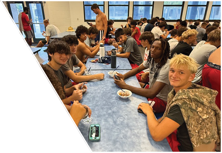 A large group of people sitting closely together at tables in a brightly lit room with large windows, some engaged in conversation and one standing while looking at a phone. Description generated by AI