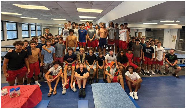 A large group of people, some wearing casual clothing and others shirtless or in athletic shorts, are gathered closely together indoors under fluorescent lighting. Description generated by AI