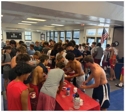 A large group of people wearing casual clothing are gathered closely around tables in a brightly lit room, engaging in an activity that involves handling small items or food. Description generated by AI