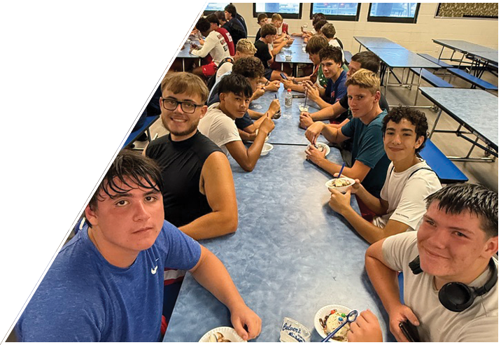 A group of people sitting at a long blue table in a cafeteria, eating from bowls and smiling at the camera. Description generated by AI