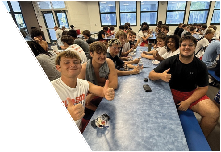 A group of people sitting at long cafeteria tables smiling and giving thumbs up in a brightly lit room with large windows. Description generated by AI