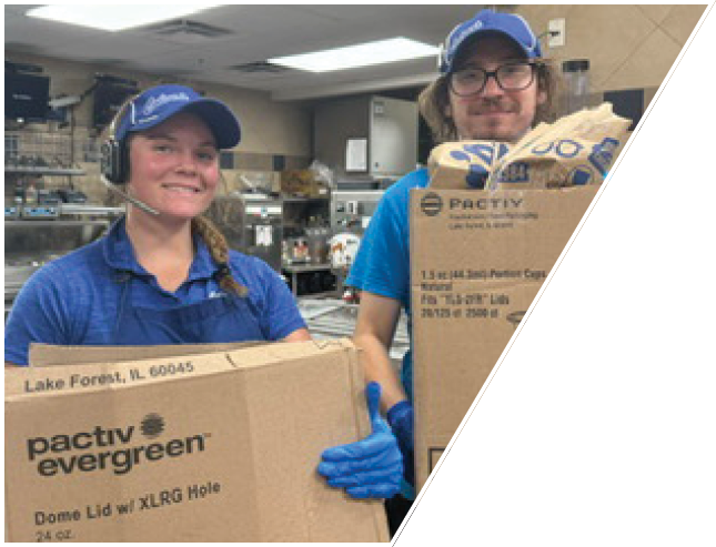 Two adults wearing blue uniforms and caps are smiling while holding large cardboard boxes in a commercial kitchen. Description generated by AI