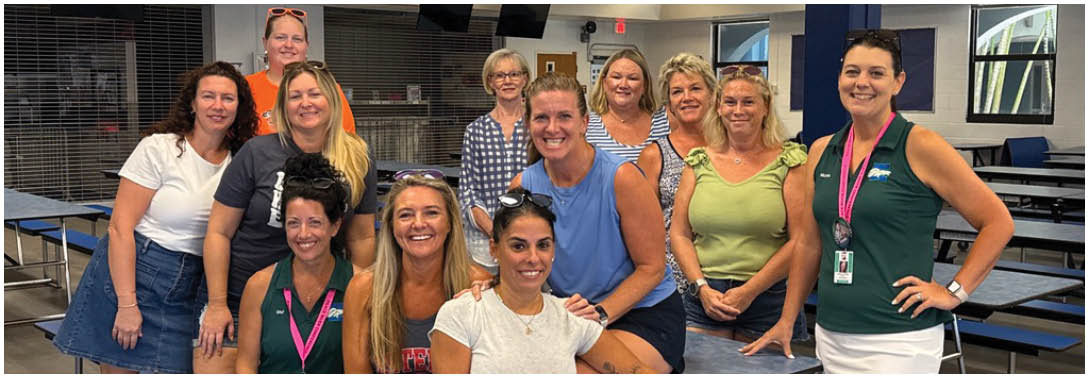 A group of adults smiling and posing together in a casual indoor setting with some sitting on and around a cafeteria-style table. Description generated by AI