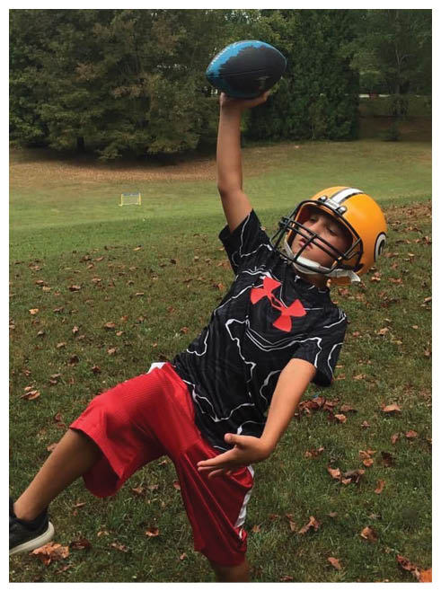 A child wearing a yellow football helmet and a black sports jersey is holding a football above their head outdoors on a grassy field with trees in the background. Description generated by AI