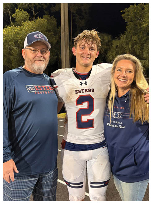 A person wearing a wet football uniform with the word Estero on it is smiling while standing next to two other people partially visible. Description generated by AI