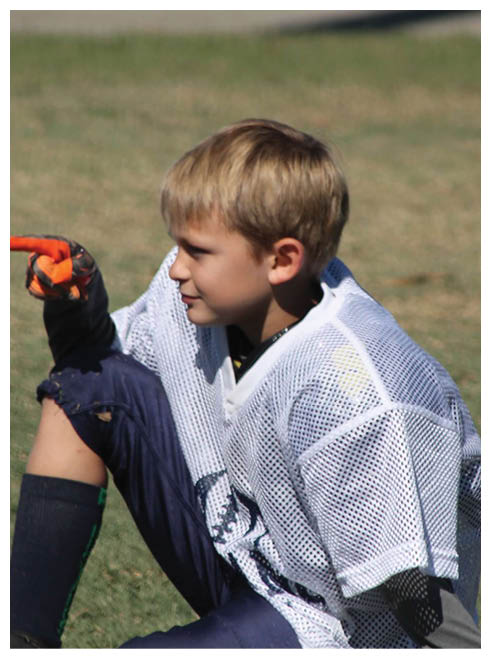 A child wearing a white mesh sports jersey, dark pants, and an orange glove is sitting on grass and pointing to the left. Description generated by AI